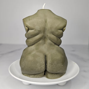 An olive green sculptural candle shaped like a stylized human torso from the back, resting on a small white pedestal plate against a light gray background.